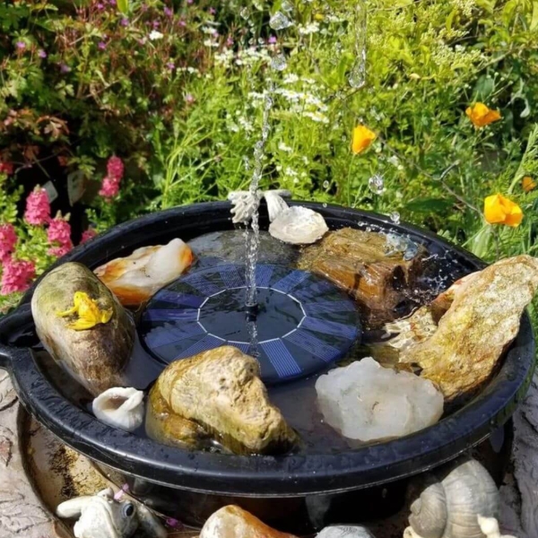 A compact garden fountain with a dark circular basin, a central solar-powered centerpiece spraying a thin stream of water, surrounded by rocks and bright flowering plants in bloom.